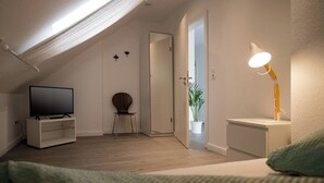 A modern bedroom with a sloped ceiling, a television, a chair, a bedside table with a lamp, and a door leading to another room. - Vacation apartment Geestland "buten un binnen" North Sea (Geestland)