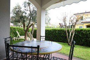 Een overdekt terras buiten, met een ronde tafel en stoelen, omringd door groen en bomen.