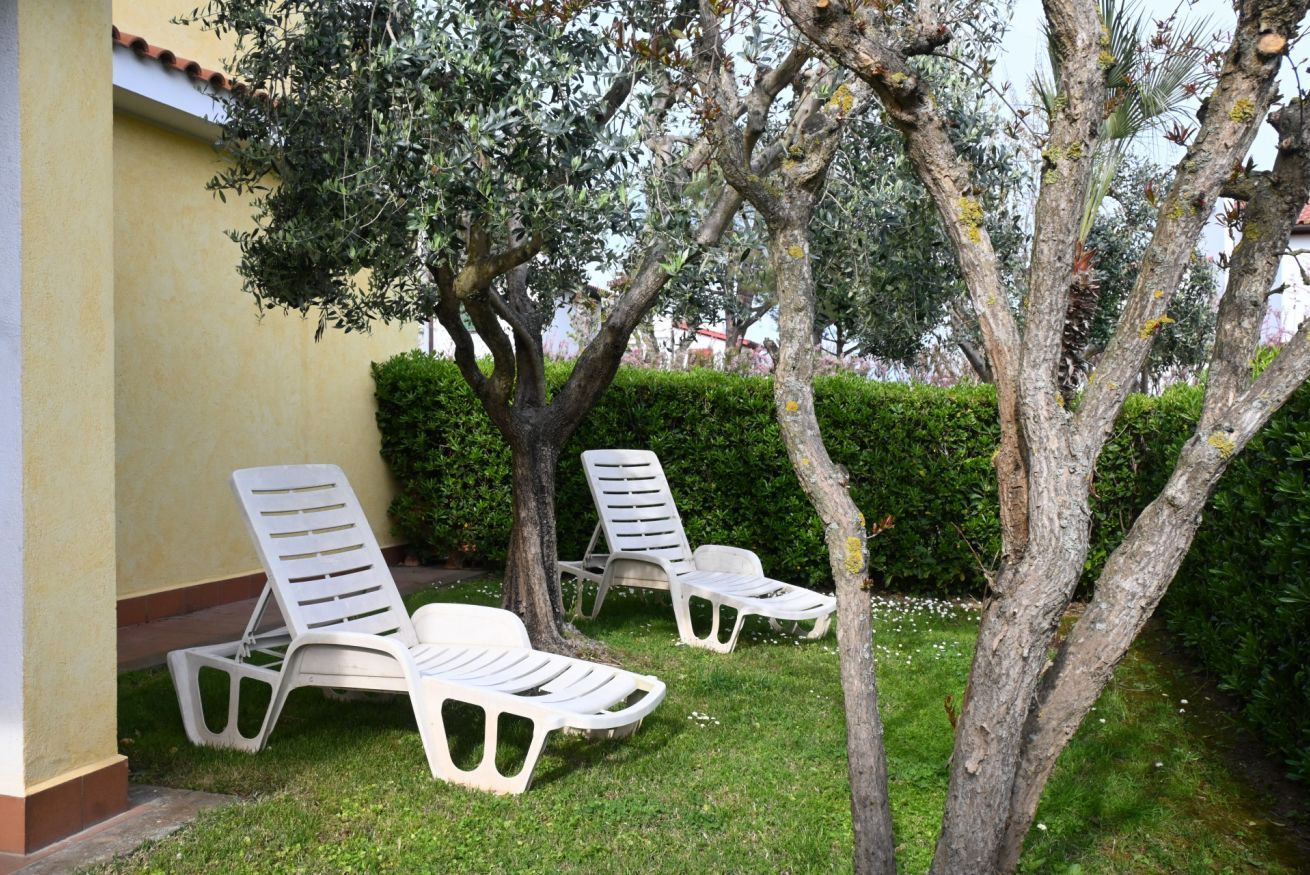 Two white lounge chairs positioned under a tree in a garden area with a hedge and a building in the background.