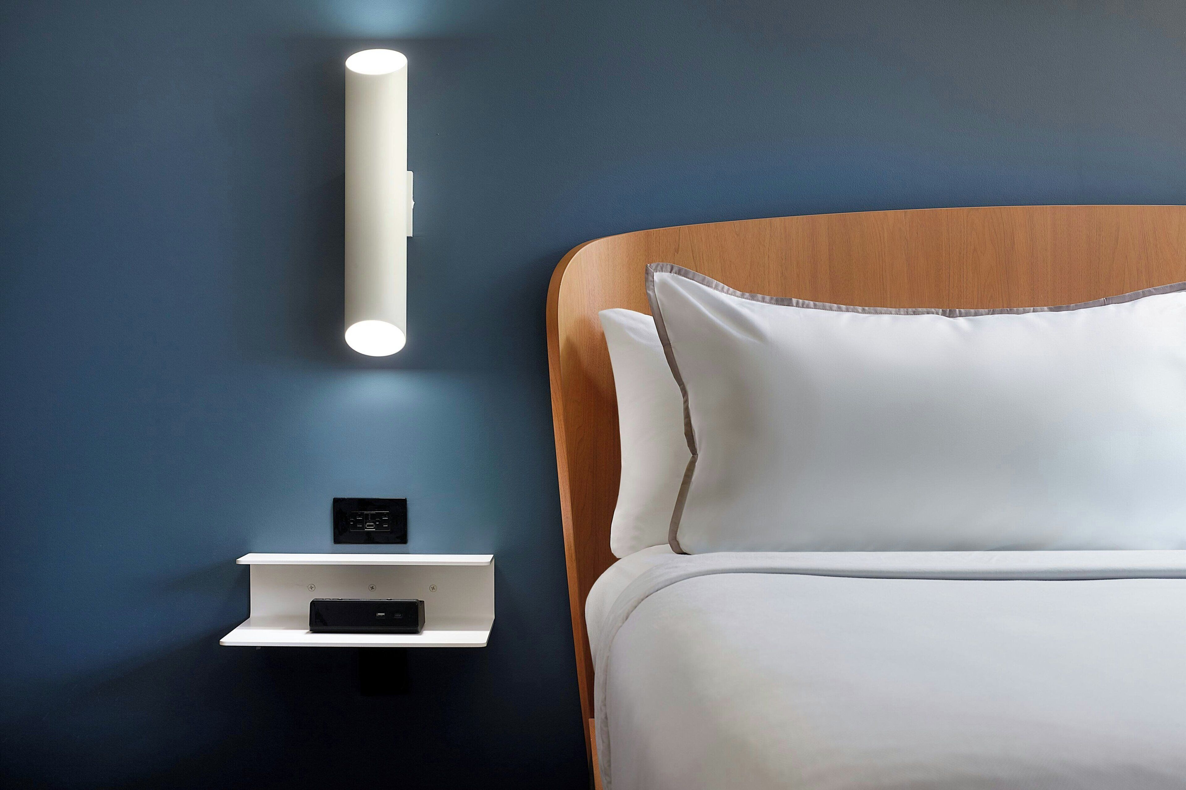A modern bedroom with a wooden headboard, white bedding, a bedside table with a power outlet, and a wall-mounted light fixture.