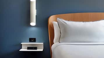 Un dormitorio moderno con cabecera de madera, ropa de cama blanca, una mesita de noche con enchufe y una lámpara fijada a la pared.