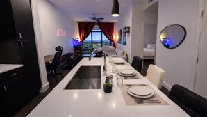 A modern kitchen with a central island, a dining area with a table set for four, and a bedroom visible through an open door. - Home Movie Theatre. Walk to FIFA/NRG/Med Center. 1BDR Highrise (Houston)