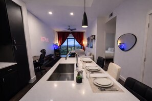 A modern kitchen with a central island, a dining area with a table set for four, and a bedroom visible through an open door. - Home Movie Theatre. Walk to FIFA/NRG/Med Center. 1BDR Highrise (Houston)