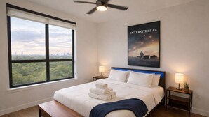 A hotel room with a large window, a bed with white linens, a blue headboard, a ceiling fan, and a poster on the wall. - Home Movie Theatre. Walk to FIFA/NRG/Med Center. 1BDR Highrise (Houston)
