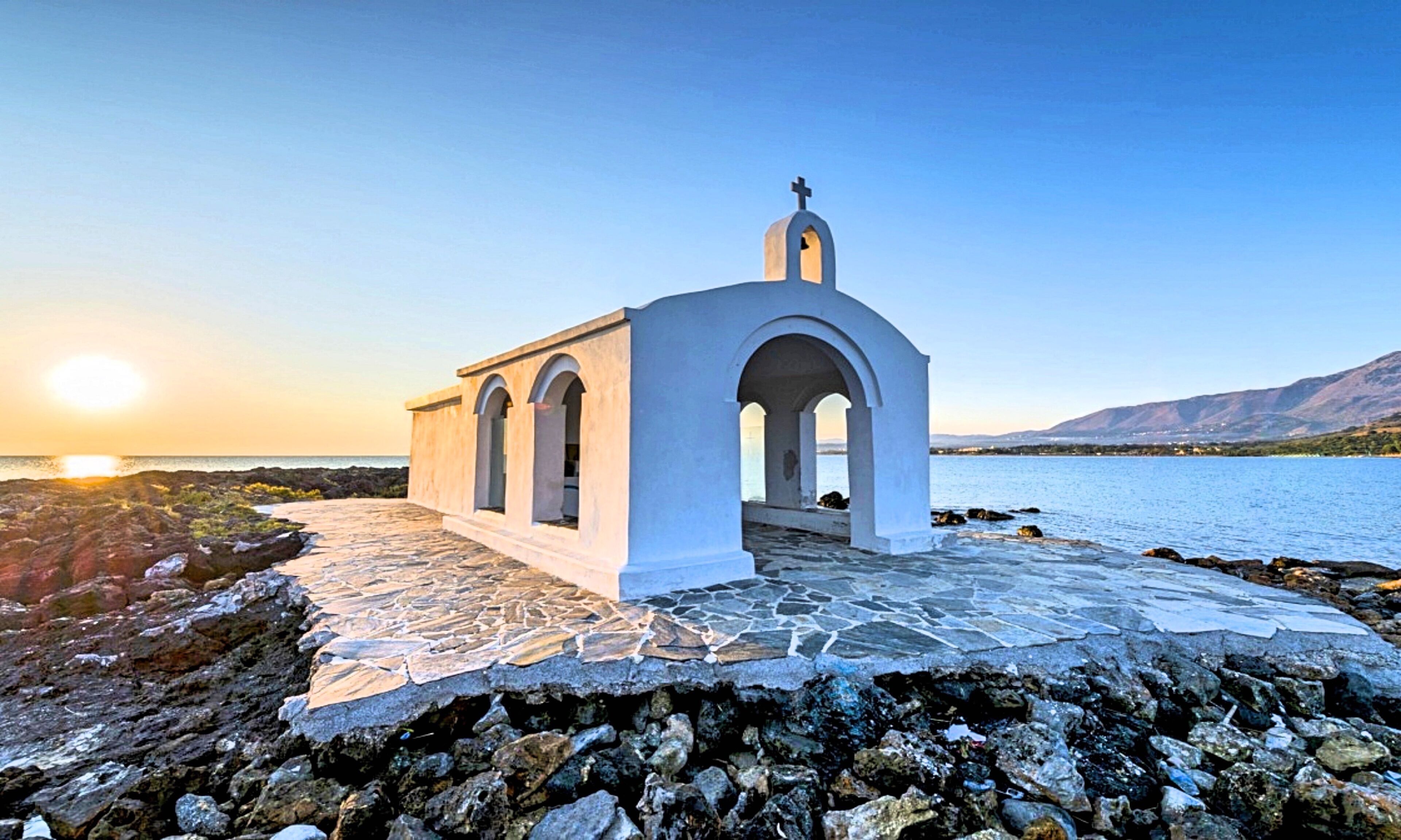 A white chapel with a cross on top, located on a rocky shore with a view of the sea and mountains in the background.