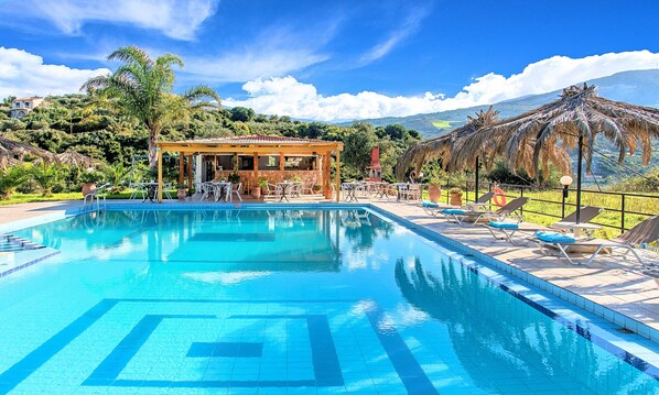 A swimming pool with lounge chairs, a covered seating area, and a mountainous backdrop.