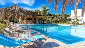 Zona junto a la piscina con sillones de descanso, un bar y una estructura con techo de paja.