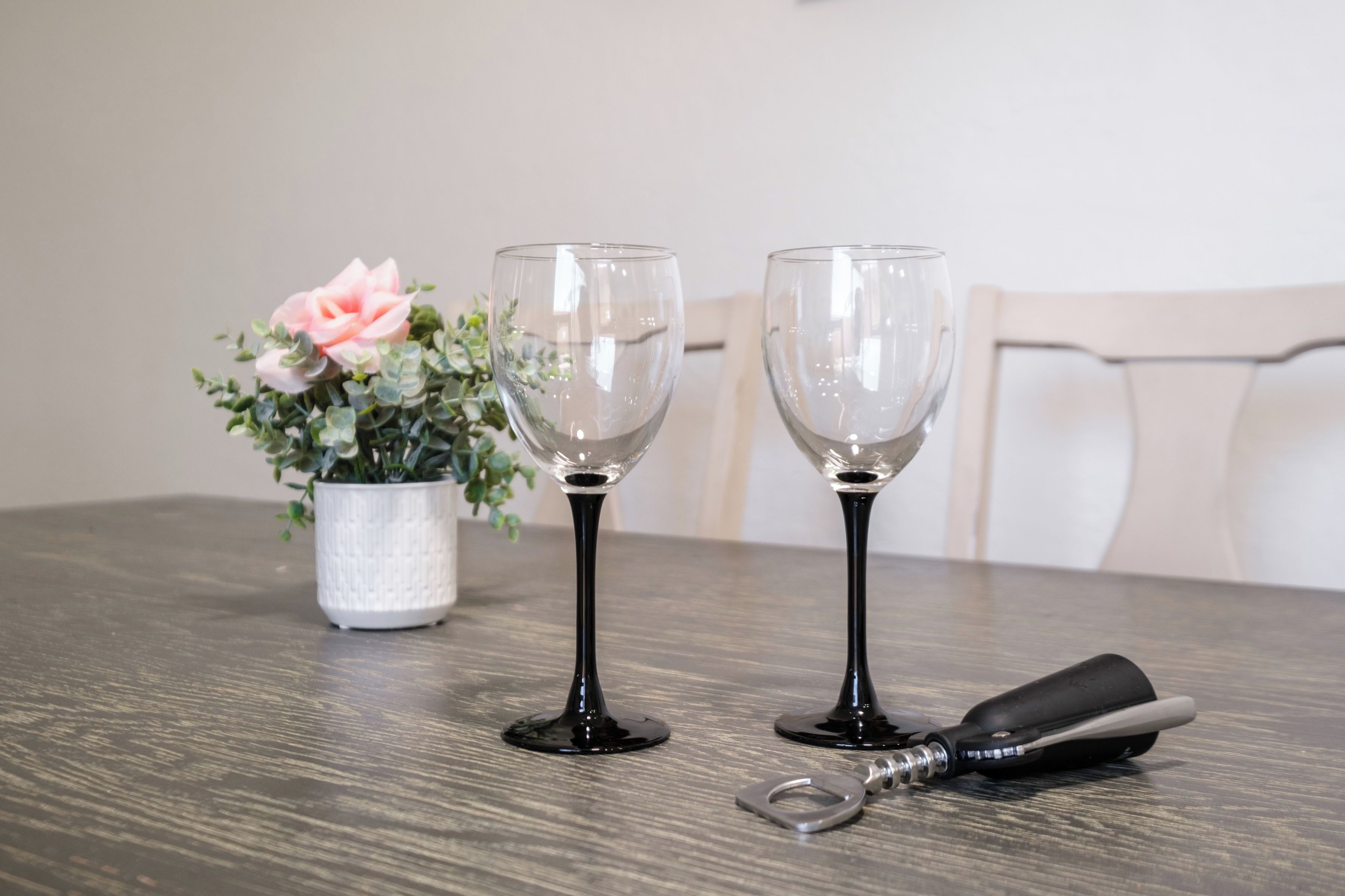 Twee wijnglazen op een houten tafel, een bloempot met een roze bloem, en een flesopener.