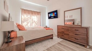 Une chambre à coucher comprenant un lit, une table de chevet, une commode, un ventilateur de plafond, un téléviseur et une fenêtre avec des rideaux.