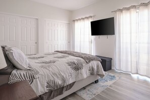 Une chambre à coucher avec un lit, un téléviseur fixé au mur, un banc et une fenêtre avec des rideaux.