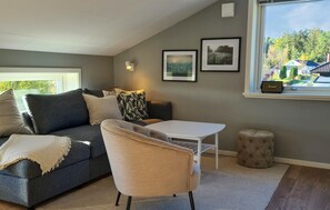 Una sala de estar moderna con un sofá gris, una mesa blanca, un sillón beige y un pequeño otomaní. Hay dos cuadros en la pared y una ventana con vistas a árboles y casas.