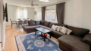 Un salon comprenant un canapé d’angle, une table basse, un tapis à motifs, un coin repas avec des chaises et un téléviseur fixé au mur.
