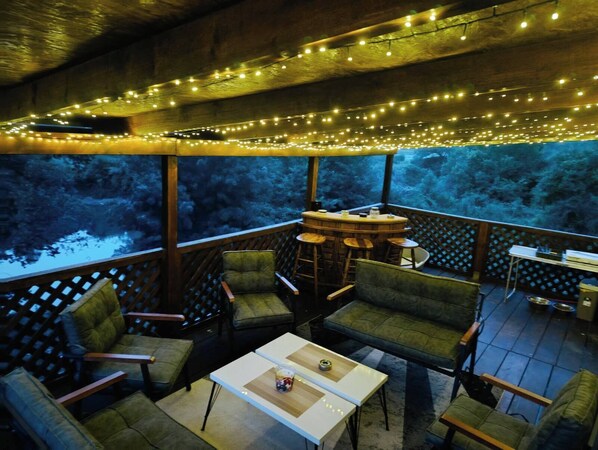 Un espace extérieur aménagé avec des chaises et des tables, des guirlandes lumineuses suspendues et une vue sur un paysage boisé.
