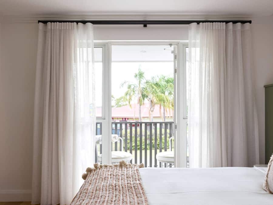 Ein Schlafzimmer mit Bett, weißen Vorhängen und Blick auf einen Balkon mit Stühlen und Palmen.