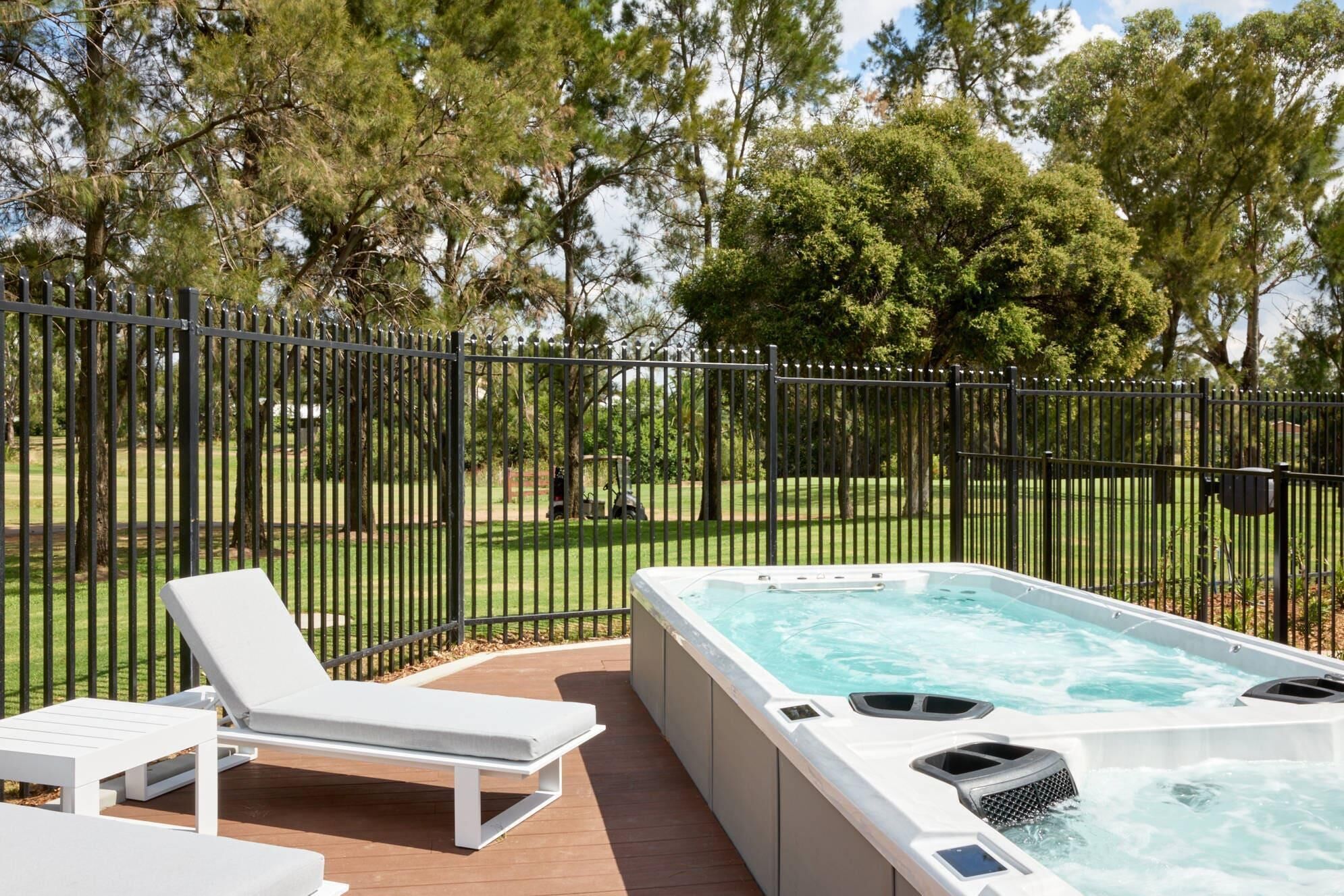 A hot tub on a wooden deck with a white lounge chair, surrounded by a black metal fence and trees.