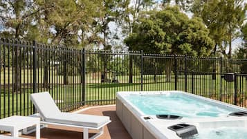 A hot tub on a wooden deck with a white lounge chair, surrounded by a black metal fence and trees.