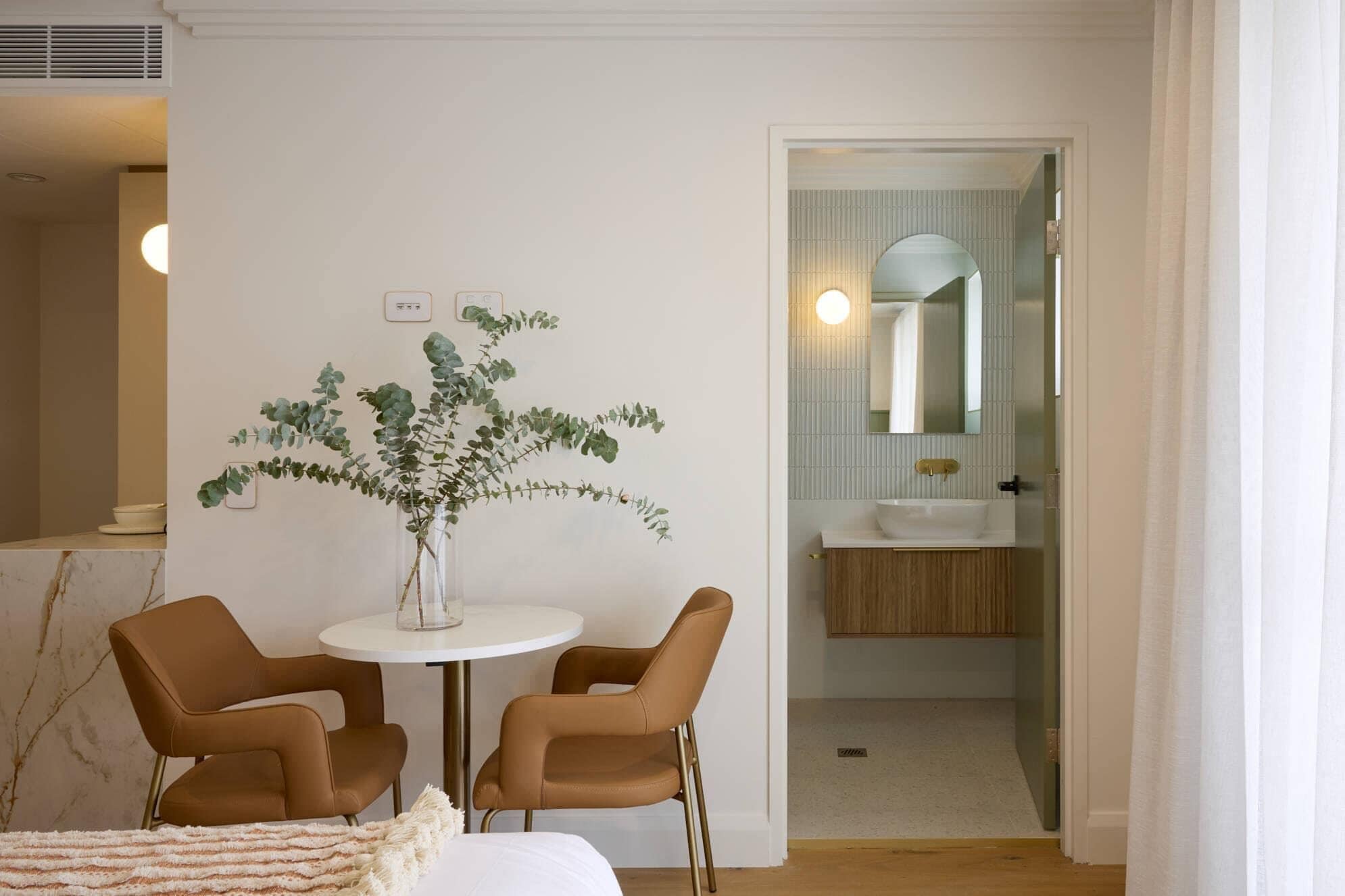 Una habitación con una mesa redonda, dos sillas, un florero con plantas y un baño visible a través de una puerta abierta.