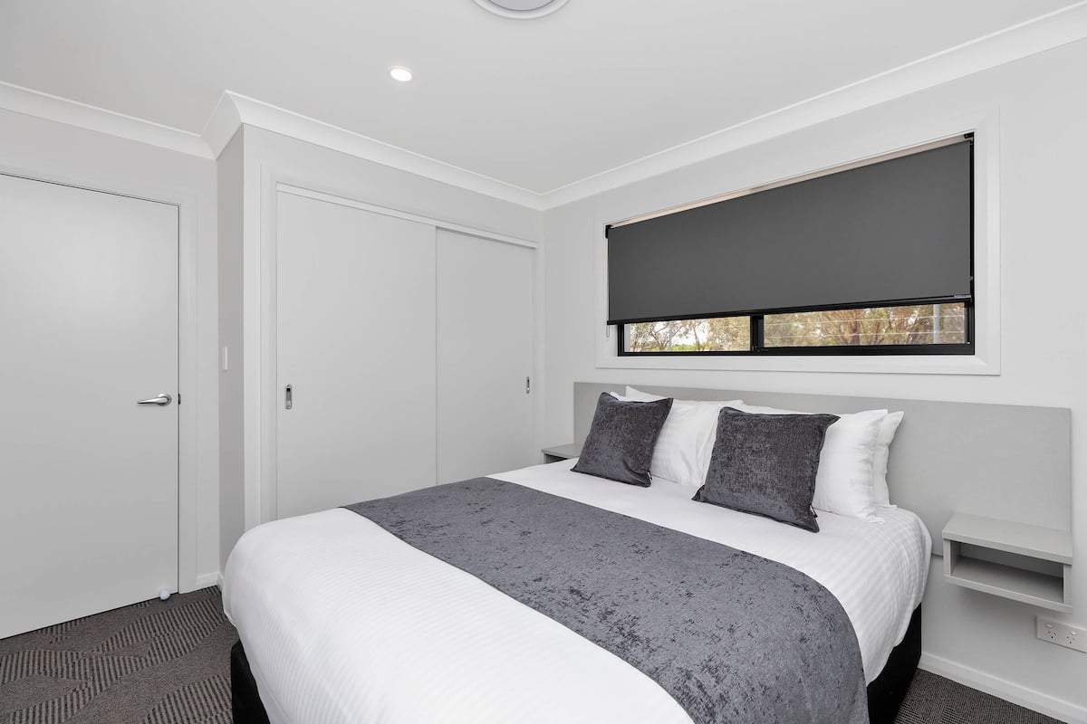 Ein modernes Hotelzimmer mit einem großen Bett, einem Fenster mit Jalousie, einer weißen Tür und einem kleinen Nachttisch.