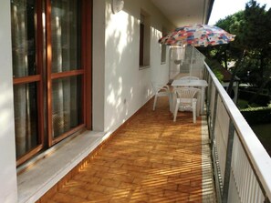 Een overdekt balkon met een houten vloer, witte stoelen en een kleurrijke parasol.
