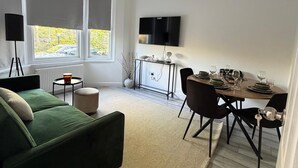 A modern living room with a green sofa, a dining table set for four, and a TV mounted on the wall. - Modern 4BR Home | Walk to Station | Sleeps 9
Minute from Tottenham Stadium (London)