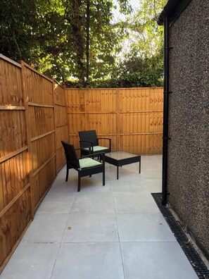 A patio with a wooden fence, a table, two chairs, and a stone wall. - Modern 4BR Home | Walk to Station | Sleeps 9
Minute from Tottenham Stadium (London)