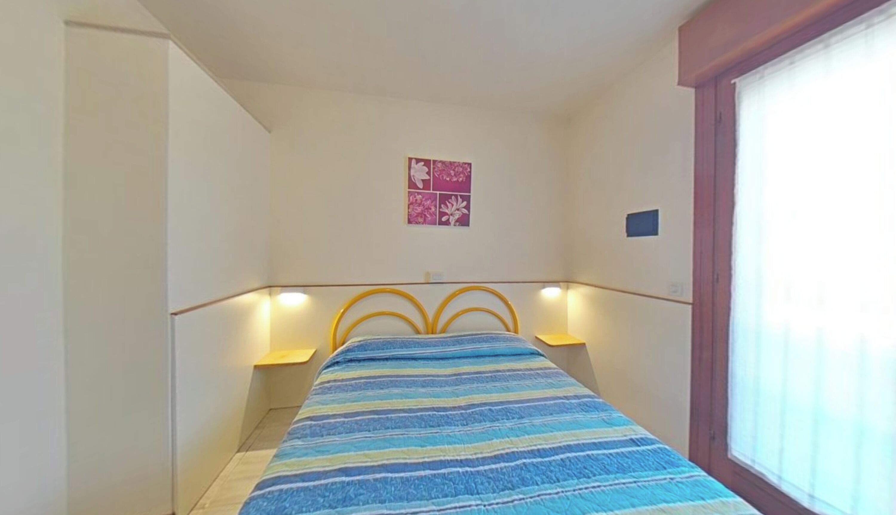 Una camera da letto con un letto, due lampade da comodino, un piccolo decoro a parete e una finestra con persiane.