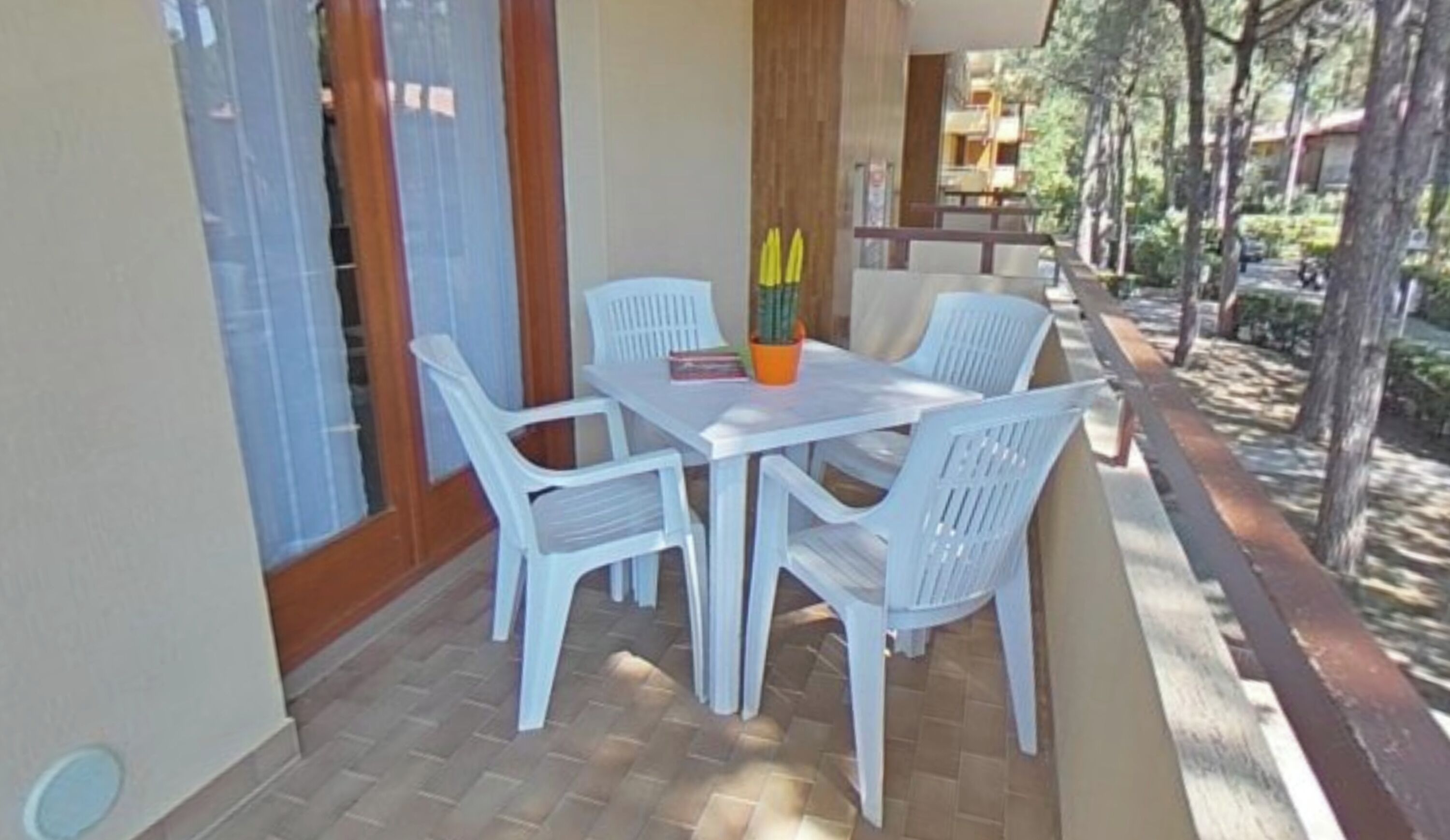 Eine Terrasse mit weißem Tisch und Stühlen, einem Blumentopf und Blick auf Bäume und Gebäude.