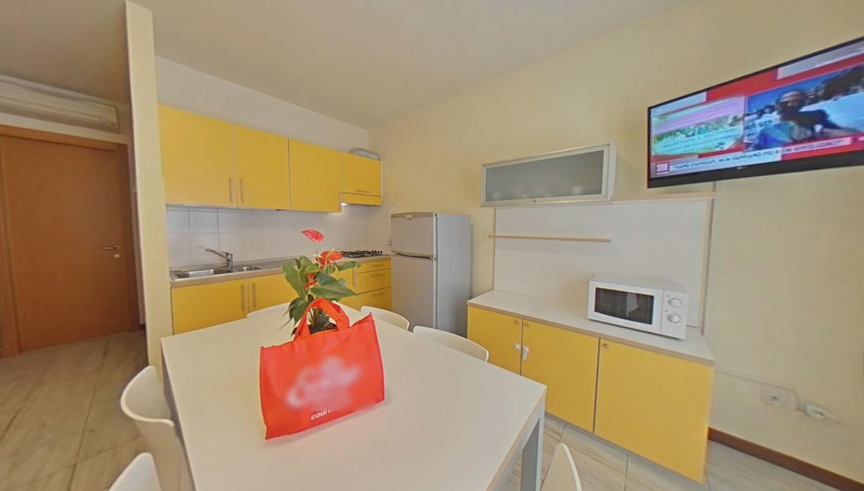 A compact kitchen with yellow cabinets, a white refrigerator, a microwave, and a dining table with a red bag.