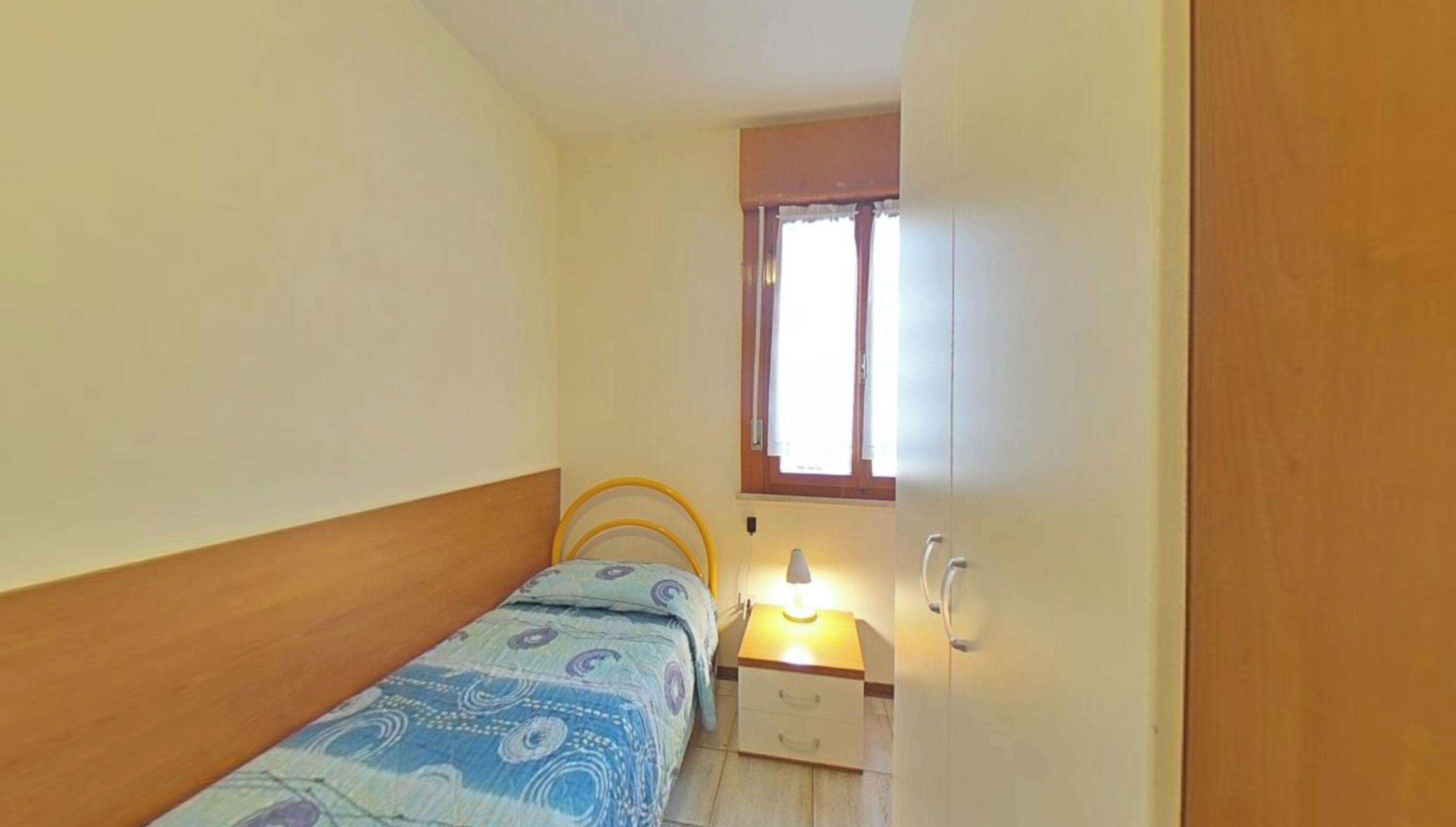 Une petite chambre avec un lit simple, une table de chevet avec une lampe, une fenêtre avec des rideaux et une armoire en bois.