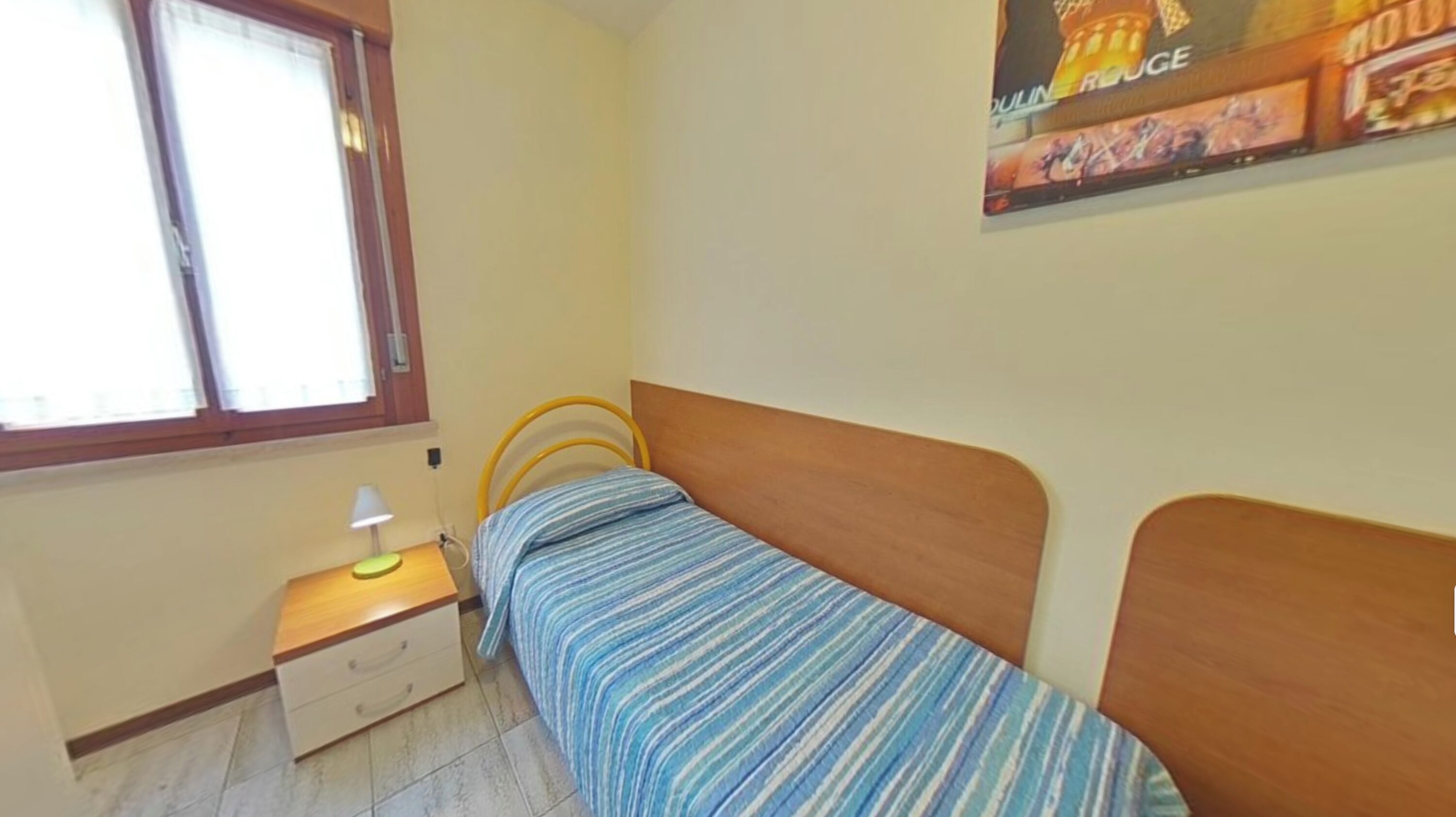 Ein kleines Schlafzimmer mit einem Einzelbett, einem Nachttisch, einer Lampe, einem Fenster und einem Poster an der Wand.