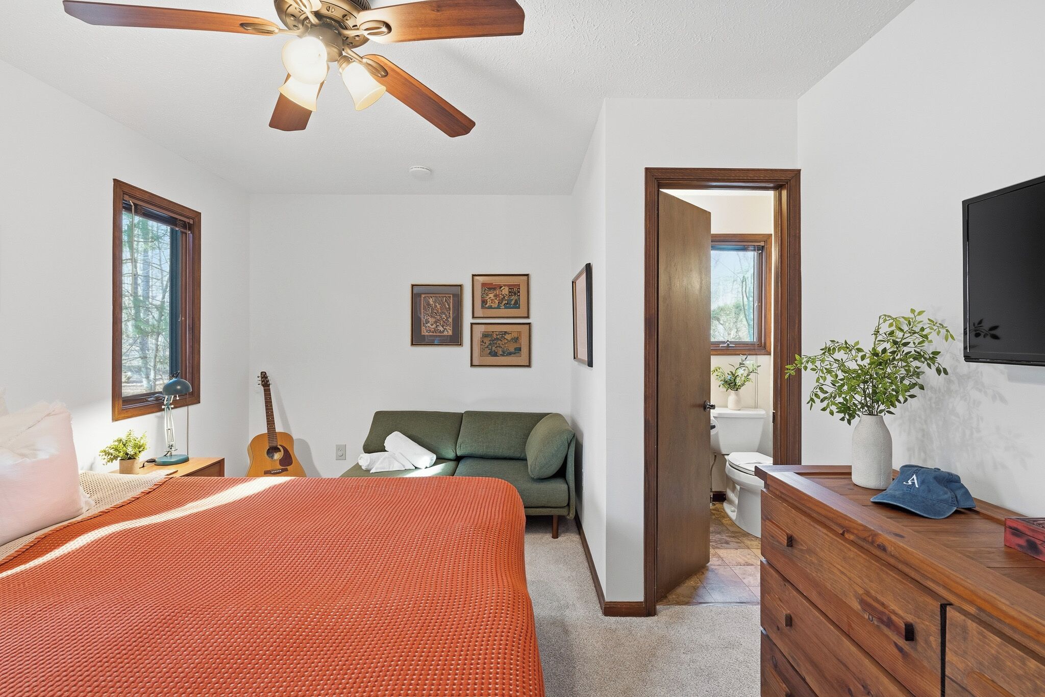 Ein Schlafzimmer mit einem Bett, einem Deckenventilator, einem Fenster, einer Gitarre, einem grünen Sofa, einem Schminktisch und einer Pflanze.