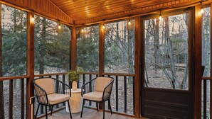 Un terrazzo in legno con due sedie e un tavolo, circondato da ampie finestre che si affacciano su una foresta.