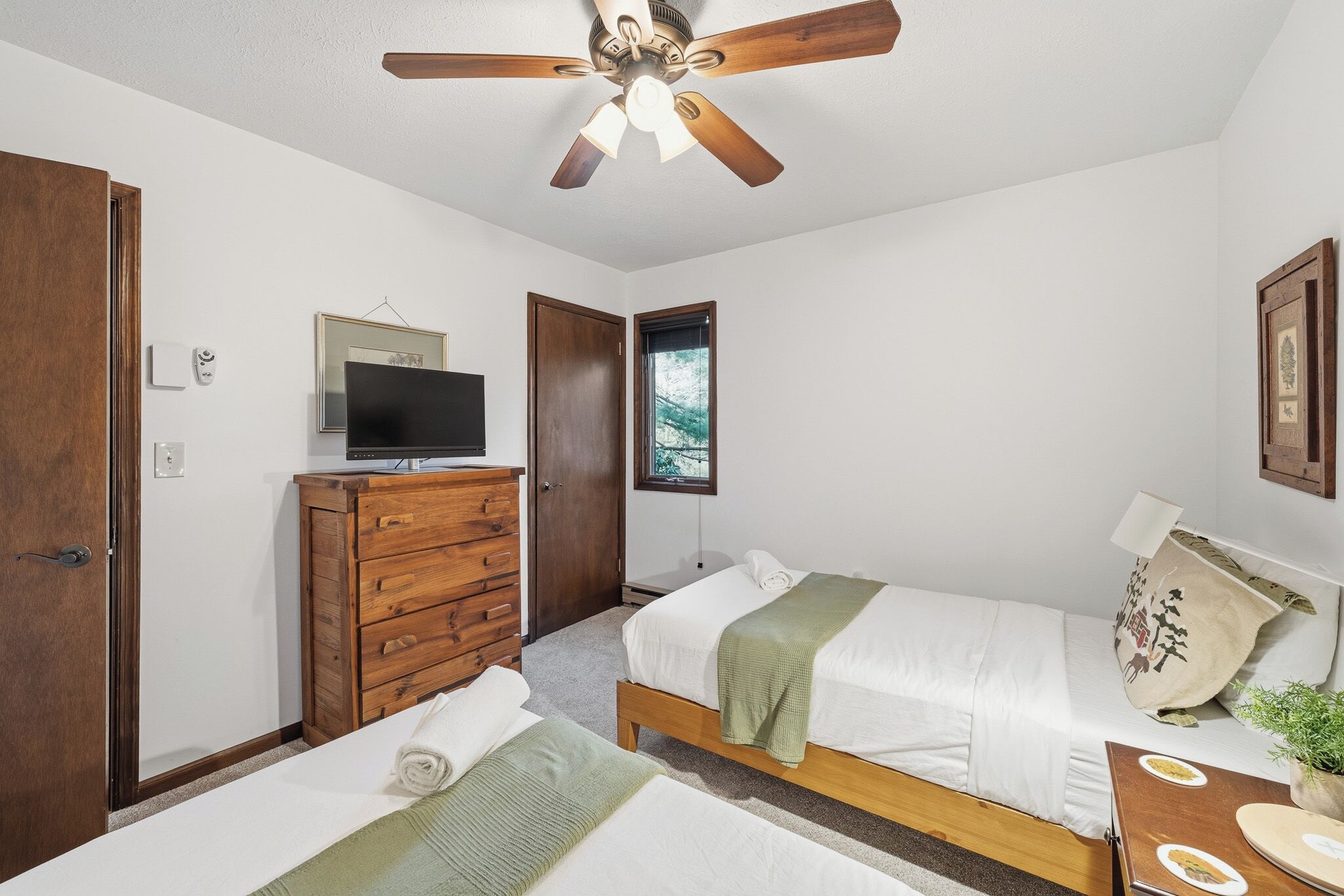 Quarto com duas camas, um ventilador de teto, uma TV, cômoda e uma janela.
