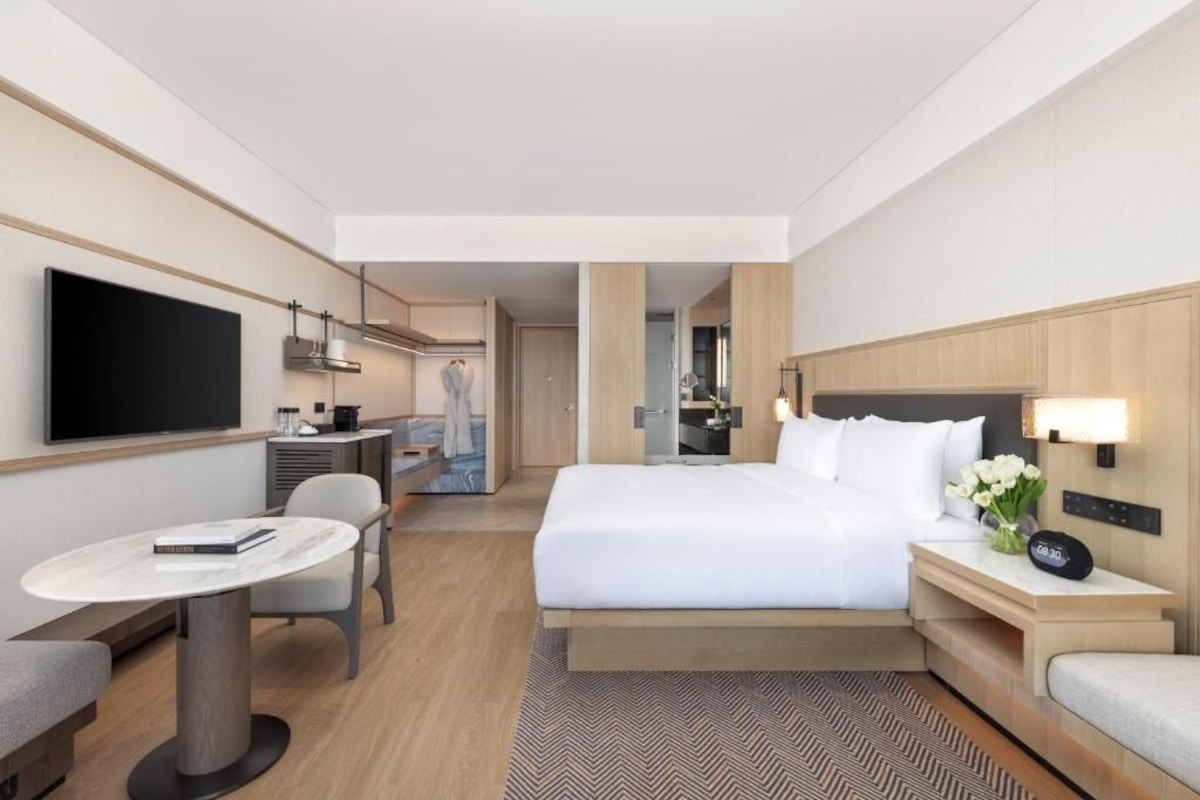 Quarto de hotel com cama, mesa de cabeceira, televisão, escrivaninha, cadeira e vista para a cozinha.