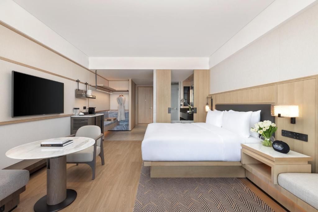 A hotel room with a bed, bedside table, TV, desk, chair, and a view into the kitchen area.