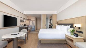 A hotel room with a bed, bedside table, TV, desk, chair, and a view into the kitchen area.