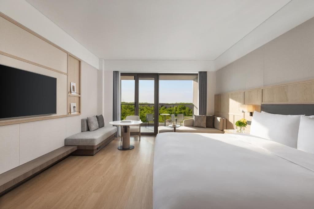 A modern hotel room with a large bed, a seating area with a table, and a balcony with a view.