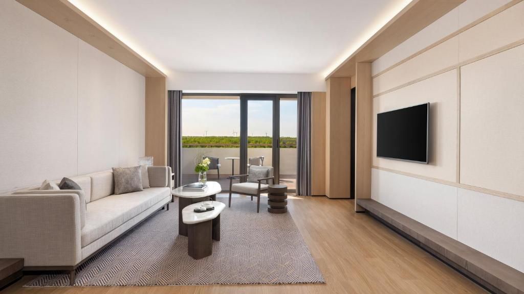 A modern living room with a sofa, coffee table, and chairs, a flat-screen TV mounted on the wall, and a large window offering a view of the outdoors.