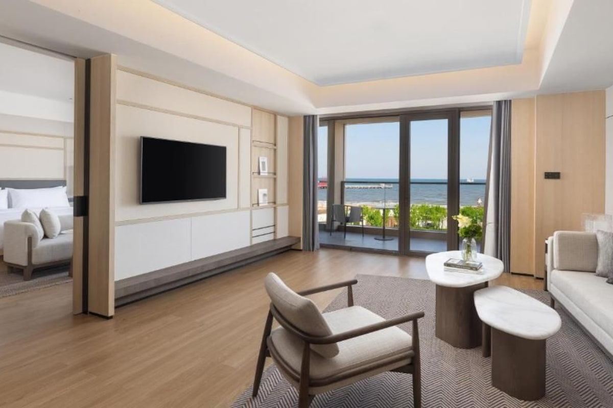 A modern hotel room with a balcony, a flat-screen TV, a sofa, a dining table, and a chair.