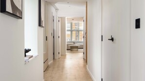 Un couloir aux murs blancs, doté d’un parquet clair, et dont les portes donnent sur une pièce avec une fenêtre et un canapé.