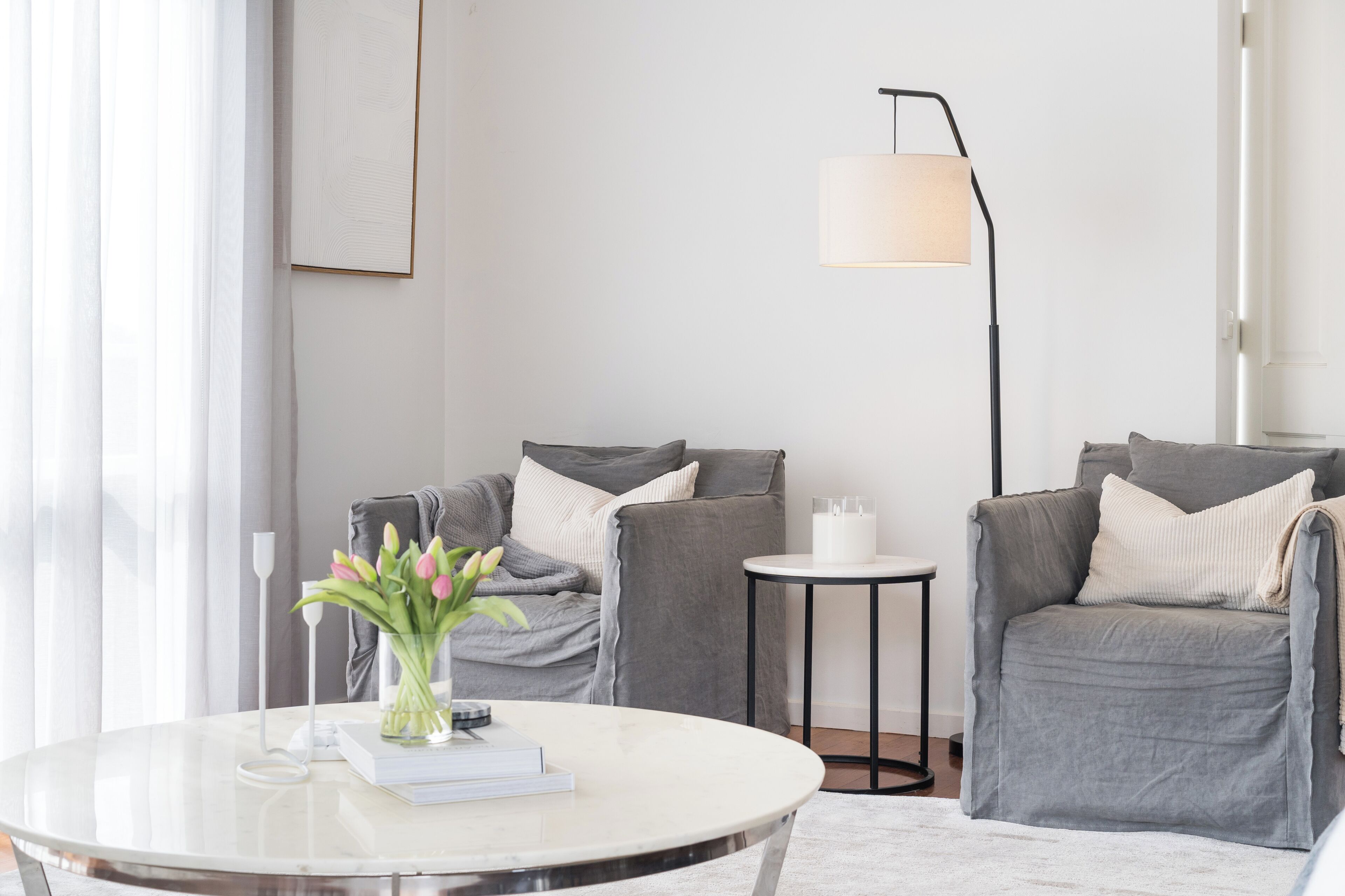 Een moderne woonkamer met een ronde witte tafel, twee grijze fauteuils, een vloerlamp en een vaas met bloemen.