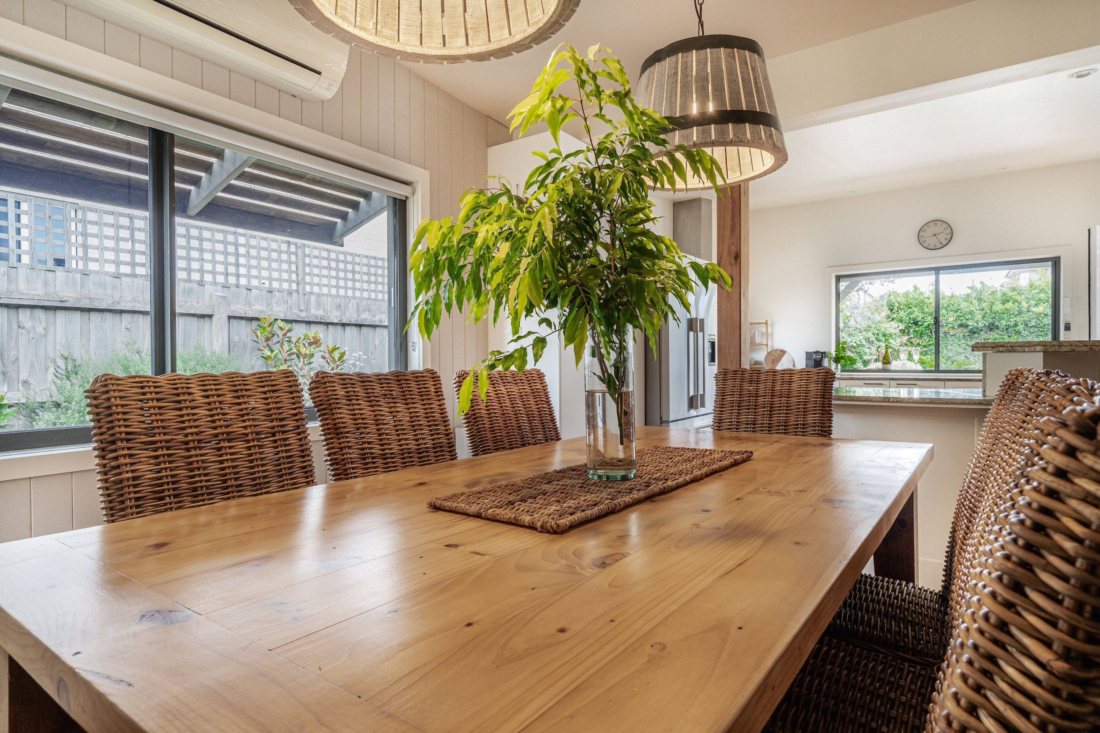 A dining area with a long wooden table, wicker chairs, a vase with a plant, and pendant lighting.
