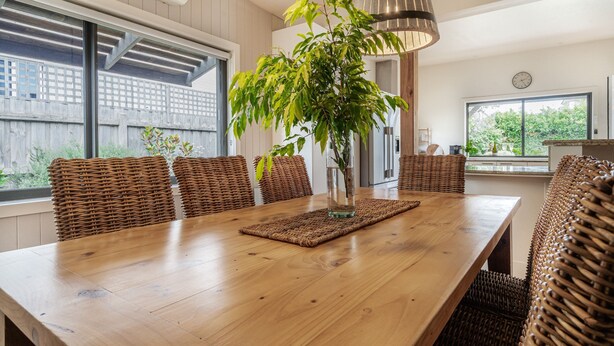 Uma sala de jantar com uma mesa de madeira comprida, cadeiras de vime, um vaso com planta e iluminação pendente.