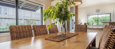 A dining area with a long wooden table, wicker chairs, a vase with a plant, and pendant lighting.