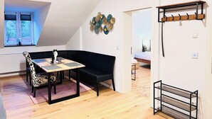 Una habitación con un sofá negro, una mesa con una silla estampada, una lámpara de techo, un adorno en la pared y una cama en la habitación contigua.