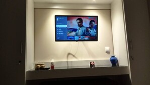 En väggmonterad tv som visar en scen från en film, en liten hylla med en vas och en flaska samt en dörr med en ljusströmbrytare.