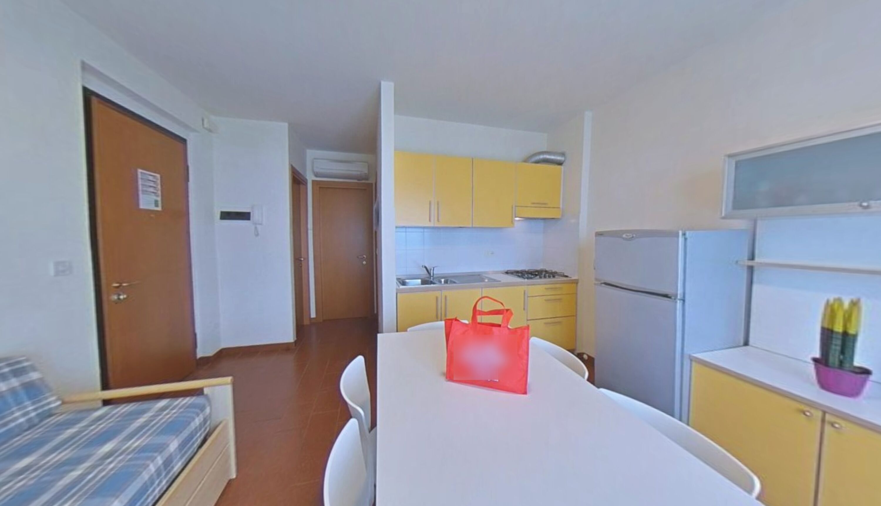 Una stanza compatta con un letto, un tavolo con una borsa rossa, un angolo cottura con mobili gialli e un frigorifero.