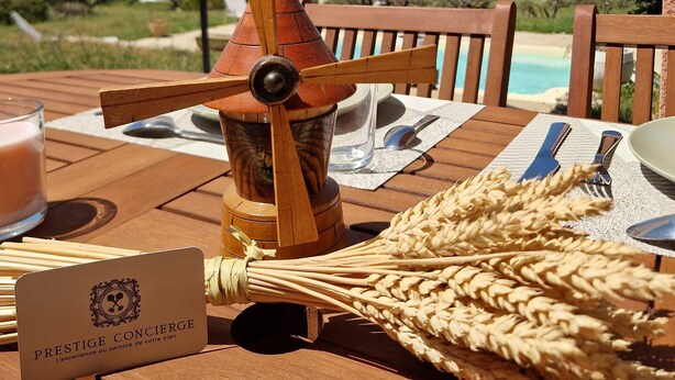 A wooden table with a sign, a windmill, a glass of liquid, and a woven basket. - Villa Oliveraie - Olive Grove Experience (Villecroze)