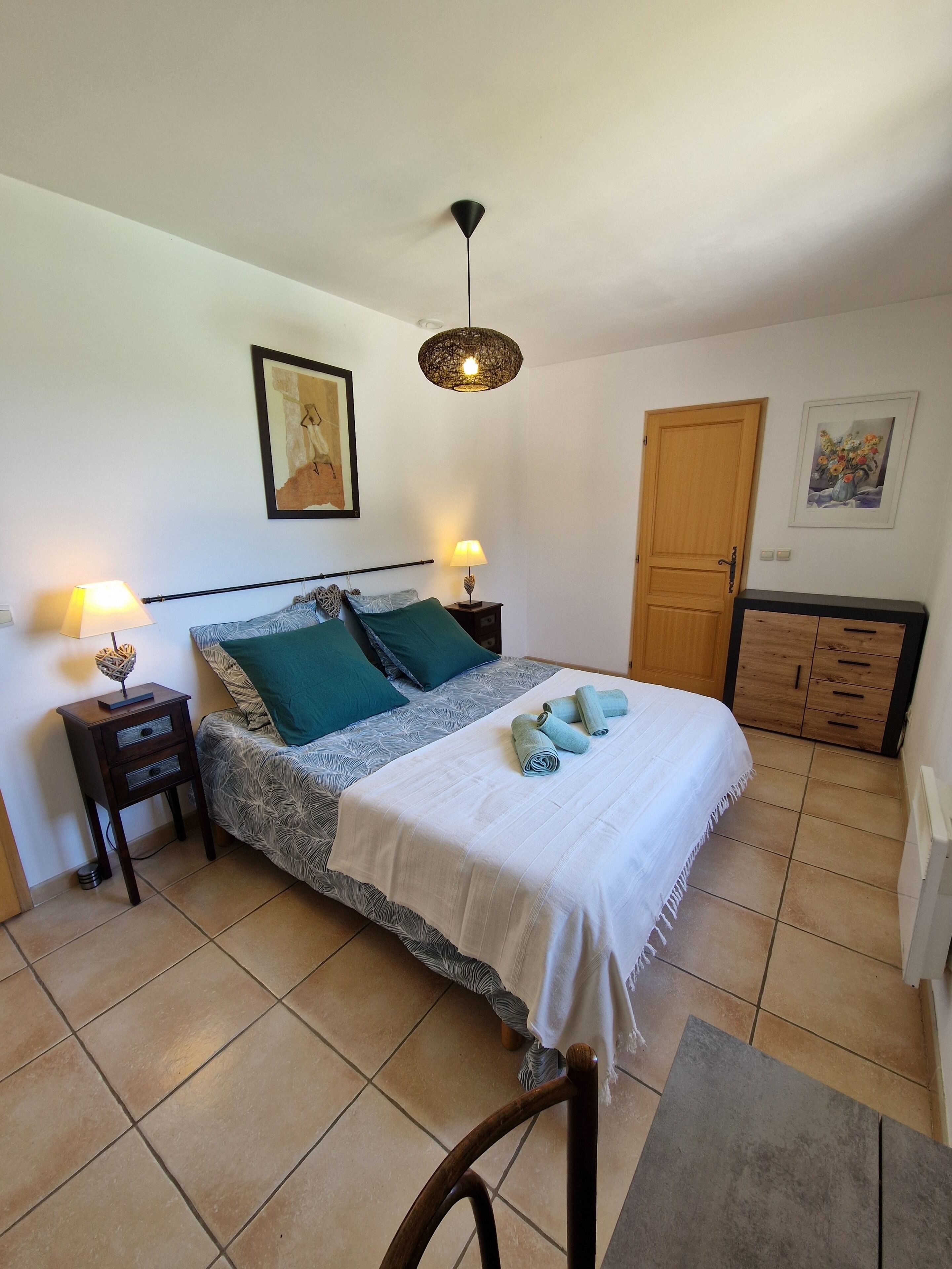 A bedroom with a bed, two bedside tables, a nightstand, a wooden wardrobe, a hanging light fixture, and a framed picture on the wall.