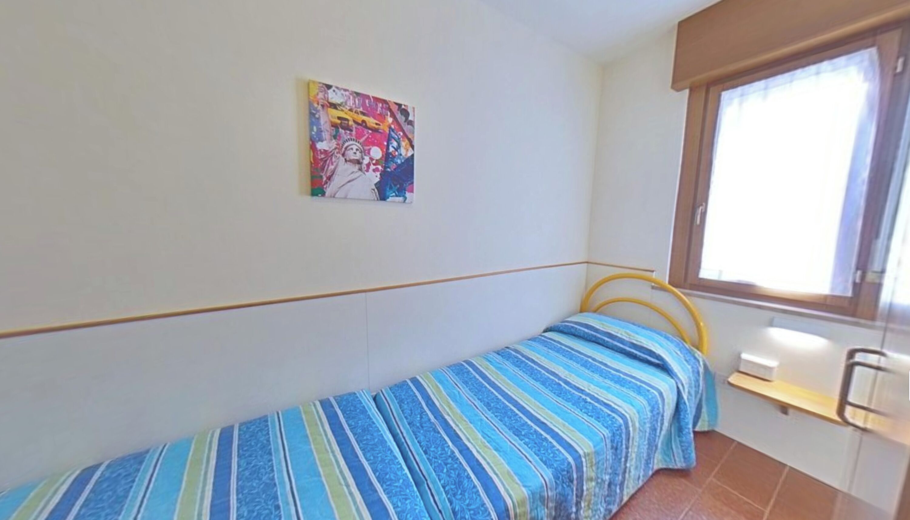 A small, single-bed room with a striped bedspread, a window with a wooden frame, and a colorful painting on the wall.